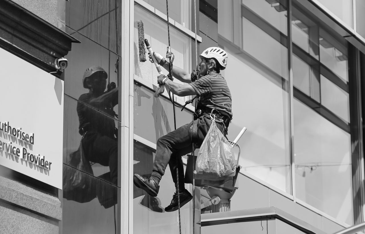 Noir et blanc vu d’en bas d’un homme cordiste, casque de protection sur la tête et équipé de matériel professionnel, nettoyant la façade vitrée d’une tour moderne